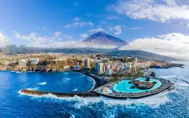 Tenerife’s coast taken from the ocean, with dark blue water, a bright blue pool, a rocky coastline, scattered buildings and a mountain partially covered in cloud