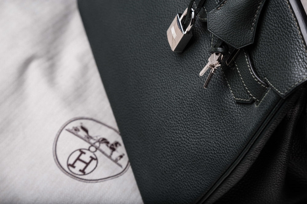 A black leather Birkin bag has a silver lock and keys, and is sitting on top of a Hermes-branded dust bag