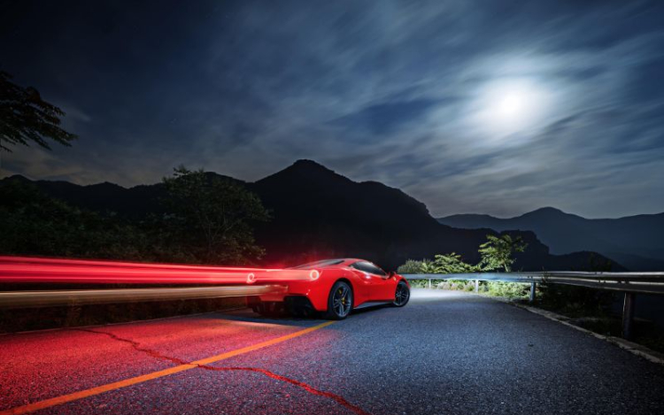 A red Ferrari driving alone on a road at night, with mountains, a silver railing, trees and the moon visible in the distance. There is a trail of red light coming from the car’s tail lights