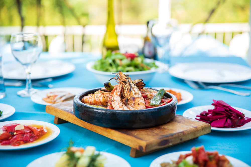 A blue table featuring a wooden board with a black bowl of shrimp, plates of seafood, appetizers and salads with a wine glass and bottle blurred in the background