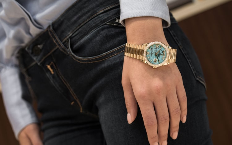 A gold Rolex watch with a light blue face on a woman’s wrist. She’s wearing a light blue shirt and dark blue denim jeans