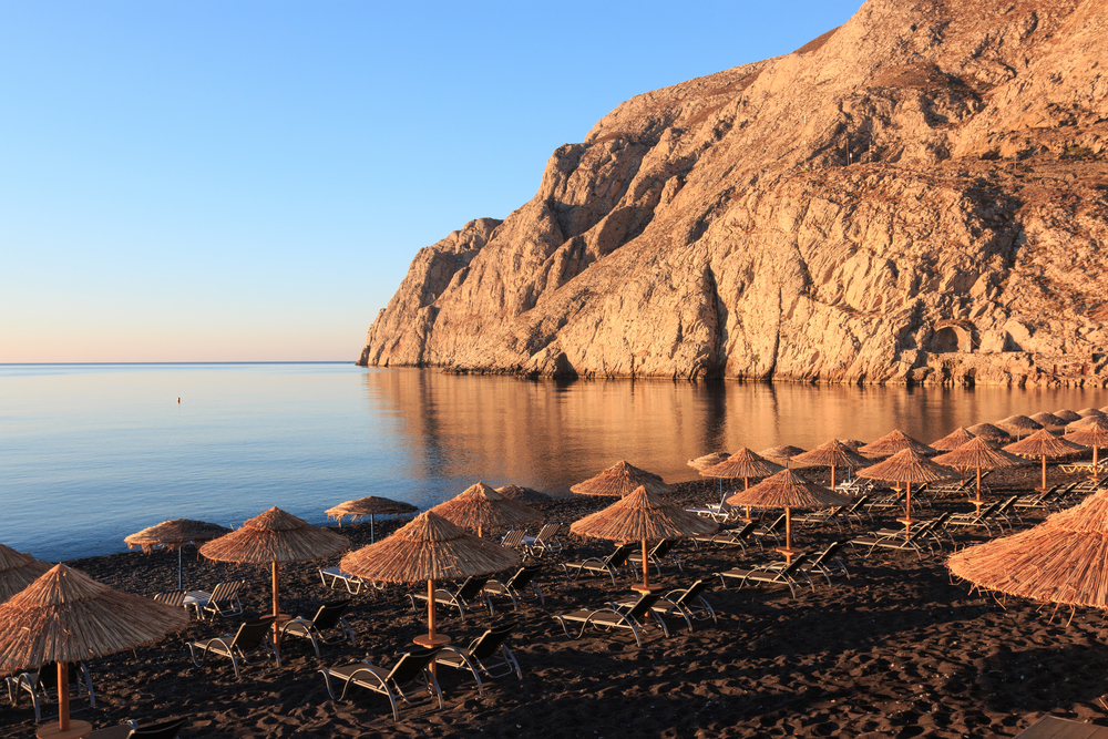 iew of Kamari beach featuring rows of brown sun loungers with umbrellas on sand with water, a big brown cliff and a sunset in the background